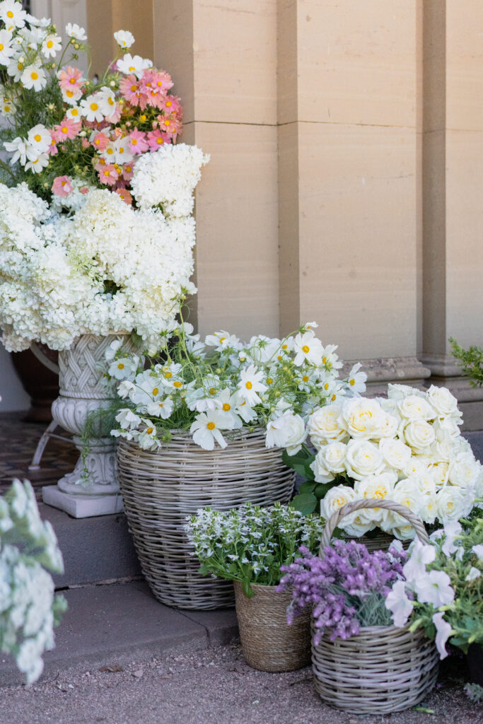 The image shows a door with flowers in front of it. There are flowers in a decorative urn and wicker baskets