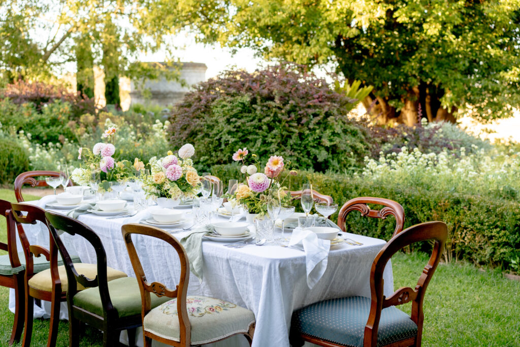 The image is of an outdoor table with chairs and flowers on it. It shows a well-set dining area with a table covered with a tablecloth and surrounded by chairs.