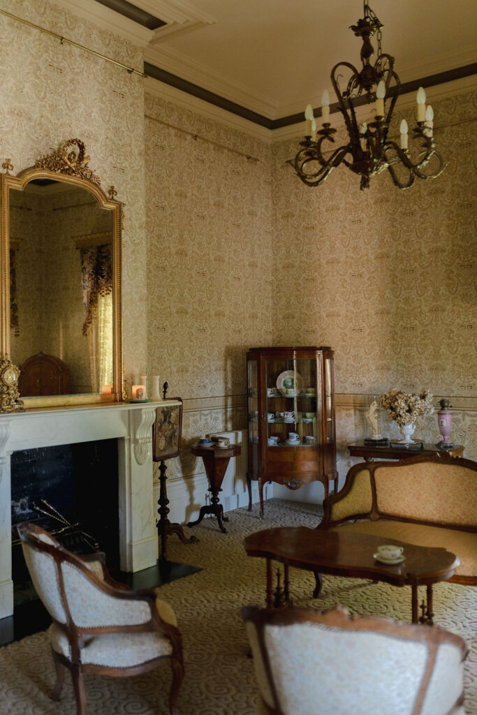 The image is a living room with a fireplace. It includes a coffee table, mirror, couch, chair, and other antique furniture.